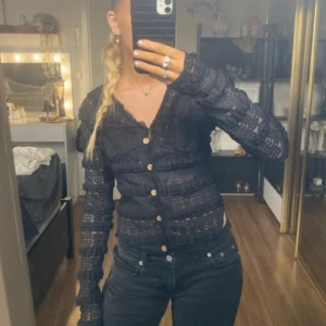 Svart spetskofta med knappar - Säljer en elegant svart spetskofta med fina träknappar. Den är långärmad och har en tight passform som framhäver figuren. Perfekt för höst och vår, och funkar både till vardags och fest. Koftan är i mycket bra skick och ger en stilren look.