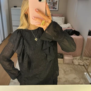 Svart glittrig blus med volangdetaljer - Säljer en elegant svart blus med glittrigt tyg och volangdetaljer på framsidan. Blusen har långa ärmar och en normal passform, perfekt för festliga tillfällen. Den är både stilren och trendig, och passar bra till både byxor och kjol.