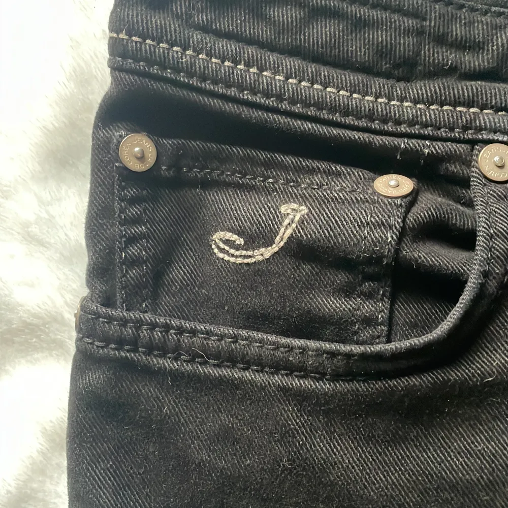 Säljer ett par snygga svarta jeans från Jacob Cohen. De har en klassisk femficksdesign med en stilren broderad detalj på myntfickan. Jeansen har knappar i gylfen och en läderpatch bak. Perfekta för både vardag och fest!. Jeans & Byxor.