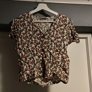 Blommig blus från Dobber - Säljer en söt blommig blus från Dobber i storlek XS. Den har korta ärmar och ett härligt blommönster i rött och grönt på vit bakgrund. Perfekt för vår och sommar! 🌸