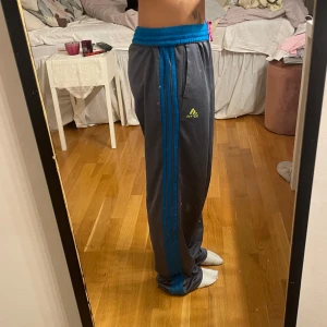 Gråa adidas byxor med blå ränder - Säljer ett par grå mjukisbyxor från Adidas med blå ränder längs sidorna. De har en elastisk midja för extra komfort. Byxorna är i bra skick💞