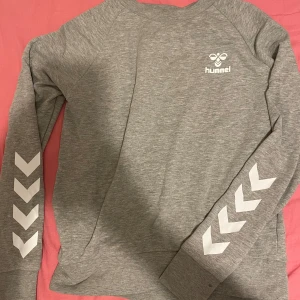 Grå sweatshirt från Hummel - Säljer en grå sweatshirt från Hummel i bra skick. Den har långa ärmar med de klassiska Hummel-pilarna i vitt längs ärmarna. Perfekt för en avslappnad stil eller träning. Superbekväm och stilren! Passar både killar och tjejer, priser går alltid att diskutera🤩Passar även Dam M