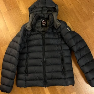 Mörklblå dunjacka från Colmar - Säljer en snygg svart dunjacka från Colmar i mycket bra skick. Jackan har en dragkedja framtill och en mysig huva. Perfekt för kalla vinterdagar! Tyvärr en skada på insidan av armen på jackan. Som tur syns de inte då de är på insidan av armen. 