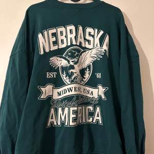 Säljer en cool grön sweatshirt med stort Nebraska-tryck på framsidan från H&M. Tröjan har en avslappnad passform och långa ärmar, perfekt för en chill dag. Materialet känns mjukt och skönt mot huden. Passar perfekt till jeans eller leggings för en casual look.