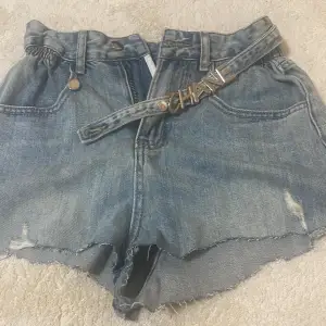 Säljer ett par coola jeansshorts i blått med slitna detaljer och ett snyggt bälte med text. Perfekta för sommaren och festivaler! De har en hög midja och knappstängning framtill. Passar perfekt till en avslappnad stil.