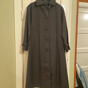 Svart kappa från Dancoat - Säljer en stilren svart kappa i trenchcoatstil från Dancoat of Denmark. Den är i mycket bra skick och har långa ärmar samt knappar och sidstöllda fickor framtill. Gjord av 65% polyester och 35% bomull med nylonfoder, superfin kvalitet. 112 cm lång Perfekt för sommar, höst och vår!