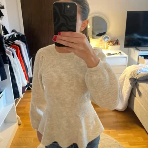 Beige stickad tröja med volangdetalj - Säljer en superfin beige stickad tröja från vero Moda. Den har en snygg volangdetalj i midjan som ger en feminin touch. Tröjan är långärmad och perfekt för höst och vinter. Den är i mycket bra skick och passar både till vardags och fest. Perfekt att matcha med jeans eller kjol!