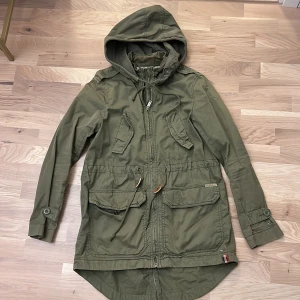 Grön parkajacka från Hilfiger Denim - Säljer en snygg grön parkajacka från Hilfiger Denim i storlek XS. Jackan har en praktisk huva och flera fickor med knappar. Perfekt för höst och vår, med en dragkedja framtill och justerbara ärmslut. Superbra skick och redo för nya äventyr! 🍂