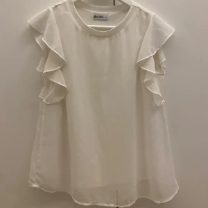 Vit blus från Miss Milo - Säljer en söt vit blus från Miss Milo i storlek S. Blusen har volangdetaljer längs ärmarna och är ärmlös, vilket gör den perfekt för sommardagar. Materialet är lätt och luftigt, vilket ger en bekväm passform. Passar både till vardags och festliga tillfällen.