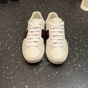 Vita sneakers från Gucci - Säljer ett par stilrena vita sneakers från Gucci i skinn. De har klassiska ränder i rött och blått på sidan och svart detalj vid hälen. Skorna är i mycket bra skick och perfekta för både vardag och fest. Storlek 41. 