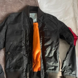 Svart bomberjacka från Alpha Industries - Säljer en klassisk svart bomberjacka från Alpha Industries i storlek small. Jackan har en snygg orange insida och är i mycket bra skick. Den har dragkedja framtill och ribbade muddar vid ärmslut och nederkant. Perfekt för höst och vår!