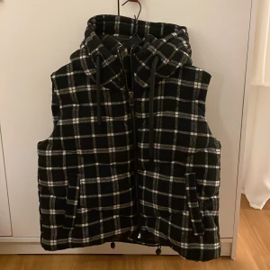 Rutig quiltad väst med dragkedja - Säljer en supermysig rutig quiltad väst. Den har en dragkedja framtill och en stor huva med snörning. Perfekt för höst och vinter när du vill hålla dig varm och snygg.