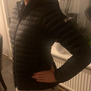 Svart dunjacka från Helly Hansen - Säljer en svart dunjacka från Helly Hansen i mycket bra skick. Jackan är perfekt för höst och vinter med sin varma och lätta design. Den har en dragkedja framtill och en huva för extra skydd mot kyla. Passformen är normal och den har långa ärmar. Perfekt för både vardag och utomhusaktiviteter!