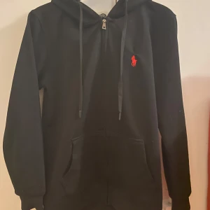 Zip hoodie Ralph Lauren - Säljer en snygg svart hoodie från Ralph Lauren med dragkedja och det klassiska röda logotypen på bröstet. Perfekt för höst och vinter. Hoodien är i nyskick eftersom att jag bara har testat den och då den var för liten för mig. Den passar om du är mellan 174-178