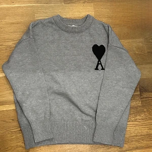 Grå stickad tröja från Ami - Säljer en snygg grå stickad tröja från Ami med deras logga i svart på bröstet. Tröjan är perfekt för både höst och vinter. Den är långärmad och har ribbade muddar vid ärmslut och nederkant. Supermysig och stilren.