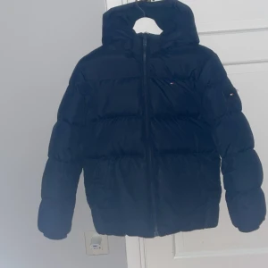 Svart pufferjacka från Tommy Hilfiger - Säljer en snygg marinblå pufferjacka från Tommy Hilfiger. Jackan har en dragkedja framtill och är perfekt för kalla dagar. Den är långärmad och har en klassisk pufferstil som håller dig varm och trendig. Perfekt för både höst och vinter!