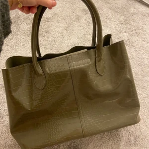 Olivgrön handväska i krokodilskinn😍 - Så fin men har bara fått användning av den en gång tyvärr. Köpt för ett år sen och endast använd en gång, så den är i nyskick fortfarande. Snygg och väldigt rymlig, den har ett fack för skoldatorn! Tillkommer ett länge band(90cm se sista bilden)💓💓