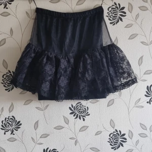 Svart spetskjol med volang - Säljer en superfin svart kjol med volangdetaljer. Kjolen är kort och har ett lager av spets som ger en elegant touch. Perfekt för festliga tillfällen eller när du vill känna dig extra fin. Den har en elastisk midja för en bekväm passform.