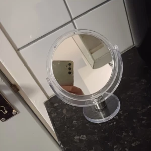 Rund sminkspegel med ställning - Säljer en praktisk och stilren rund sminkspegel med en stabil ställning. Spegeln har en genomskinlig ram och en fot i silverfärgad metall, vilket ger den en modern look. Perfekt för sminkbordet eller badrummet!