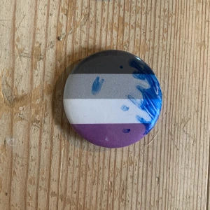 Rund knapp med pridefärger - En rund knapp med horisontella ränder i svart, grått, vitt och lila. Den har också blå färgstänk på ena sidan, vilket ger den en unik och konstnärlig touch. Perfekt för att fästa på en väska eller jacka för att visa stöd och stolthet. (Lgbtq flagga) 