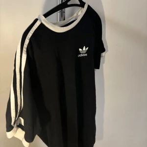 Svart och vit tröja från Adidas - Säljer en snygg svart och vit tröja från Adidas. Den har de klassiska tre ränderna på ärmarna och Adidas-loggan på bröstet. Perfekt för en sportig look eller bara för att chilla i. Nypris: 299kr