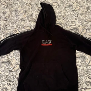 Svart hoodie från Emporio Armani - Säljer en snygg svart hoodie från Emporio Armani med EA7-logga på bröstet och längs ärmarna. Den är perfekt för en casual look och har en skön passform. Huvan har dragsko och det finns en stor ficka framtill. Perfekt för höst och vinter!