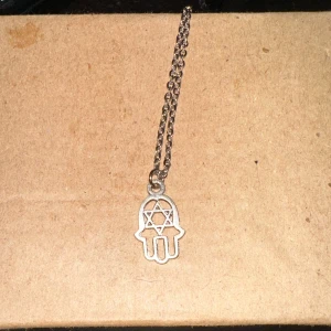 Silverfärgat halsband med hamsa-berlock - Säljer ett stilrent silverfärgat halsband med en hamsa-berlock. Berlocken har en stjärndesign i mitten och hänger på en tunn kedja. Perfekt för att ge en subtil touch till din outfit!