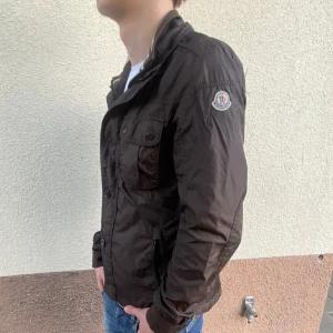 Moncler Field Jacket  - Snygg brun jacka från Moncler med knappar och en broderad logga på ärmen. Jackan har en klassisk design med flera fickor och långa ärmar. Perfekt för en stilren look.