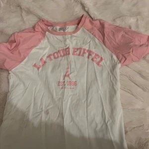 Vit och rosa t-shirt med tryck - Säljer en vit t-shirt med rosa ärmar och tryck 'La Tour Eiffel' i rosa på framsidan. Perfekt för en avslappnad stil.