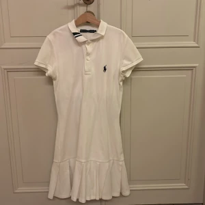 Vit tennisklänning från Ralph Lauren - Säljer en stilren vit klänning från Ralph Lauren med korta ärmar och en klassisk krage. Klänningen har en volangdetalj vid fållen och en broderad logotyp på bröstet. Perfekt för en avslappnad och elegant look.