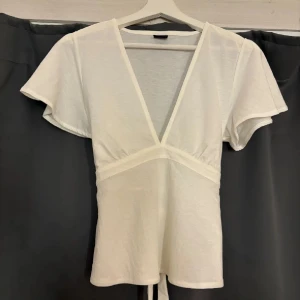 Vit v-ringad blus - Säljer en stilren vit blus med v-ringning och korta ärmar. Blusen har en knytning framtill som ger en fin detalj. Perfekt för en avslappnad men chic look.