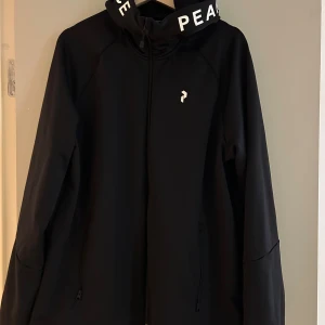 Svart jacka från Peak Performance - Säljer en stilren svart jacka från Peak Performance med dragkedja och huva. Jackan har långa ärmar och textdetaljer på huvan. Perfekt för en sportig look eller vardagsbruk.