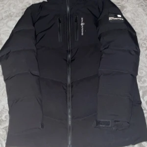 Svart dunjacka från Sail Racing - Säljer en svart dunjacka från Sail Racing med huva och dragkedja. Jackan har flera praktiska fickor med dragkedjor och en logotyp på ärmen. Perfekt för kalla dagar.