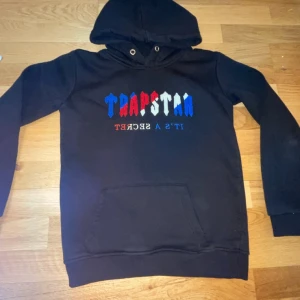 Svart hoodie med broderad text - Säljer en svart hoodie med broderad text i blått, rött och vitt. Hoodien har en klassisk design med känguruficka och justerbar huva. Perfekt för en avslappnad stil.