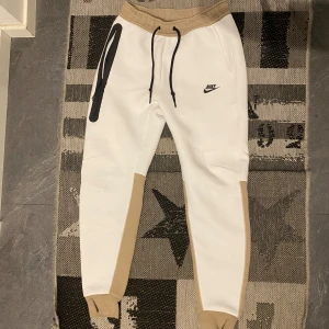 Vita och beige mjukisbyxor från Nike - Snygga vita mjukisbyxor från Nike med beigea detaljer och svart logga. Byxorna har en elastisk midja med snörning och praktiska fickor med dragkedja. Perfekta för en avslappnad stil. Dem e oanvända 