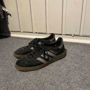 Svarta Adidas Spezial sneakers - Snygga svarta Adidas Samba sneakers i mocka med klassiska tre ränder i grått på sidan. Skorna har en brun sula och snörning framtill. Perfekta för en stilren och sportig look.