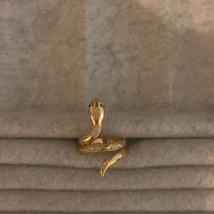 Guldfärgad ormring med stenar - Snygg guldfärgad ring formad som en orm, dekorerad med glittrande stenar längs kroppen. Perfekt för att ge en unik touch till din stil.