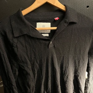 Svart pikétröja från Kronstadt - Säljer en stilren svart pikétröja från Kronstadt i extra fin merinoull. Tröjan har en klassisk krage och knappar framtill. Perfekt för en avslappnad men ändå elegant look.