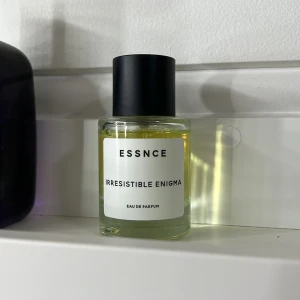 Irresistible Enigma Eau de Parfum från Essnce - Säljer en flaska Irresistible Enigma Eau de Parfum från Essnce. Parfymen kommer i en stilren, genomskinlig glasflaska med svart kork och en enkel vit etikett. Doften är mystisk och lockande, perfekt för den som vill sticka ut. Knappt använd se bild för mängd 