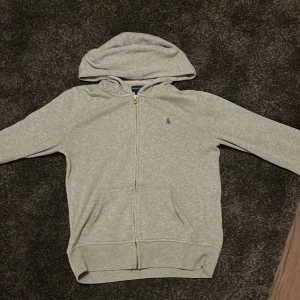 Grå hoodie från Ralph Lauren 10-12 år - Säljer en stilren grå hoodie från Ralph Lauren. Den har en dragkedja framtill och en liten logotyp på bröstet. Perfekt för en avslappnad look med sina långa ärmar och bekväma passform.