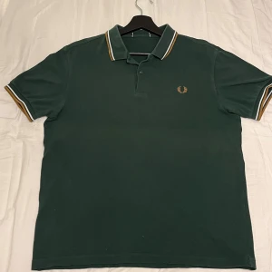 Grön pikétröja från Fred Perry - Säljer en snygg grön pikétröja från Fred Perry med klassisk krage och ränder i vitt och orange. Tröjan har korta ärmar och en broderad logotyp på bröstet.