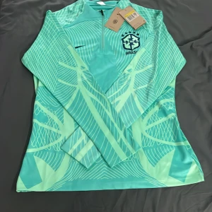 Turkos fotbollströja från Nike - Snygg turkos fotbollströja från Nike med Brasilien-emblem på bröstet. Tröjan har ett unikt mönster och en halv dragkedja framtill. Perfekt för fotbollsfans som vill sticka ut på planen.