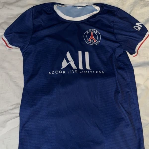 Blå PSG fotbollströja - Säljer en blå PSG fotbollströja med vit och röd detaljering. Tröjan har korta ärmar och klubbens emblem på bröstet. På baksidan finns namnet 'Mbappé' och nummer 7. 