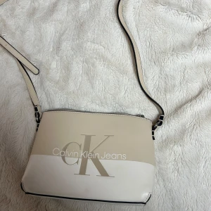 Beige och vit axelväska från Calvin Klein - Snygg axelväska från Calvin Klein i beige och vitt med justerbar axelrem. Väskan har en dragkedja upptill och det ikoniska CK-logotypen tryckt på framsidan. Perfekt för att bära dina nödvändigheter med stil.