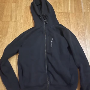 Svart hoodie från Sail Racing  - Säljer en svart hoodie från Sail Racing med dragkedja och huva. Den har långa ärmar och en stilren design med logotyp på bröstet. Perfekt för en avslappnad stil. Storlek M