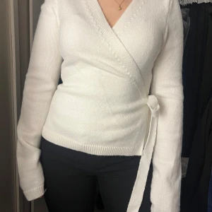 Vit omlottstickad tröja - Säljer en stilren vit omlottstickad tröja med knytband i midjan. Tröjan har långa ärmar och en v-ringad design som ger en elegant look. Perfekt för kyligare dagar.