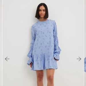 Ljusblå klänning med volangdetaljer - Charmig ljusblå klänning med blommigt mönster och volangdetaljer. Den har långa ärmar och en öppen rygg med knytband. Perfekt för en somrig look!