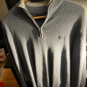 Blå stickad tröja från Lexington - Säljer en stilren blå stickad tröja från Lexington med dragkedja vid halsen. Tröjan har en diskret logotyp på bröstet och är perfekt för kyligare dagar. Den har långa ärmar och en ribbad struktur som ger en klassisk look. Finns fler bilder vid förfrågan!