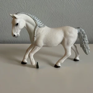 Schleich häst - Detaljerad vit hästfigur med flätad man och svans i grått. Perfekt för samlare eller som leksak för hästälskare. Hästen står i en elegant pose med lyft ben.