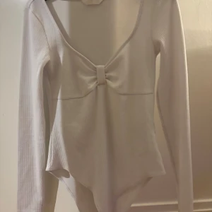 Vit ribbad body med rosett - Säljer en stilren vit ribbad body från h&m med långärmad design och en söt rosett framtill. Perfekt för en elegant och bekväm look. Bodyn har en v-ringad halslinje som ger en feminin touch. 
